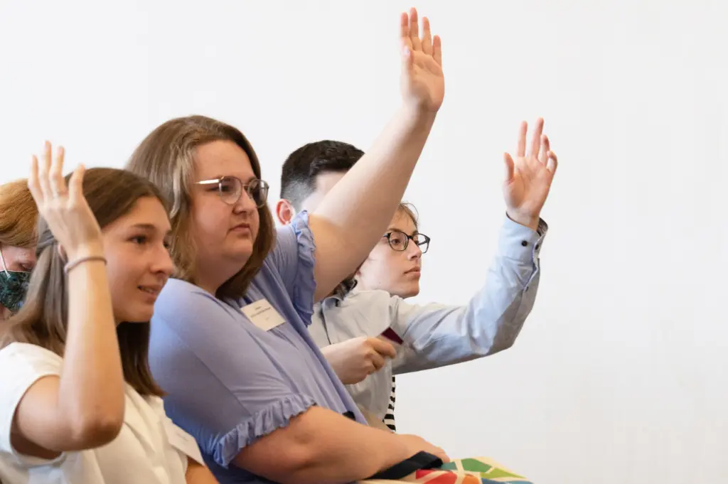 Vier Jugendliche sitzen in einer Reihe, drei heben die Hand.