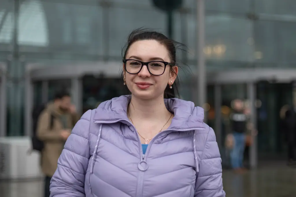 Ein Portrait der Ukrainerin, Victoria. Sie trägt eine schwarze Brille, eine lilafarbene Winterjacke und hat die schwarzen Haare zu einem Zopf nach hinten gebunden. Außerdem trägt sie eine bronzene Kette mit dem Trysub der Ukraine.