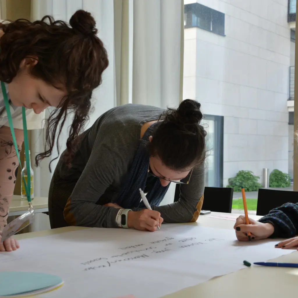 Drei junge Frauen schreiben auf einem Plakat ihre Forderungen nieder.
